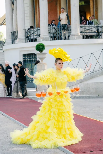 Cocktail hour entertainment with a champagne skirt server in a yellow ruffled gown, offering drinks in a stone-columned courtyard.