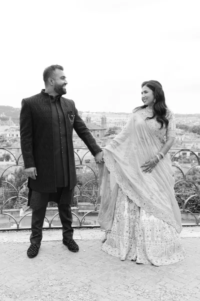 Couple portrait of a bride and groom holding hands on a terrace, the bride in a bridal lehenga, with a cityscape beyond.