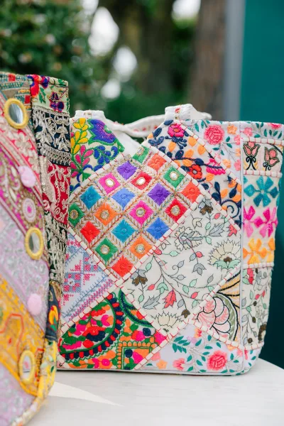 Embroidered tote bag, a patchwork handbag with mirror work, rests on a white tabletop before a teal wall and garden trees.