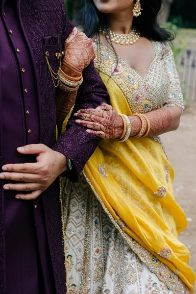 Couple portrait with the bride's bridal mehndi hands resting on the groom's sherwani as they stand on a garden path by an iron fence.
