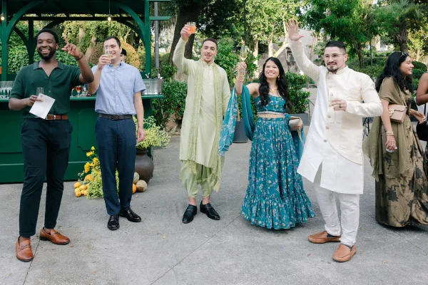 Wedding toast sparks cocktail hour cheers as guests raise champagne flutes on a garden patio under string lights among garden trees.