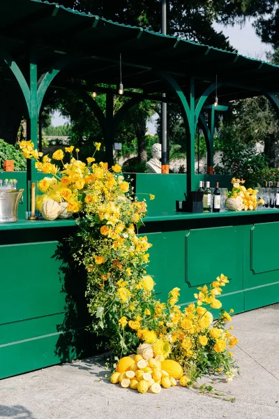 Wedding bar decor for a cocktail hour bar, accented by lemons on a green counter beneath a garden pergola with surrounding trees.