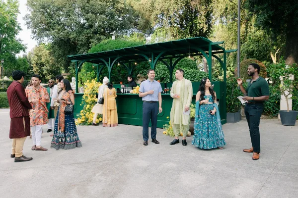 Wedding cocktail hour at an outdoor cocktail reception, guests mingle as a guest in a lehenga chats amid garden trees and hedges.