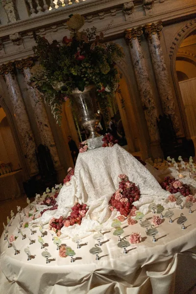 Escort card display on a floral escort card table encircling a silver urn centerpiece, set in an ornate hall with marble columns and arches.