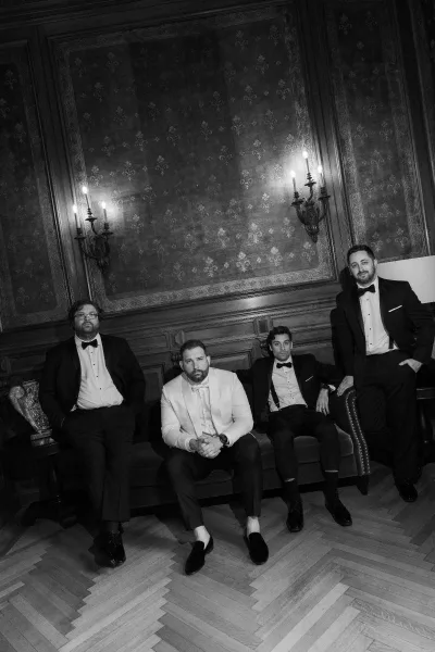 Groomsmen portrait of the groom and groomsmen, the groom in a white tuxedo jacket, in a wood‑paneled room with patterned wallpaper.