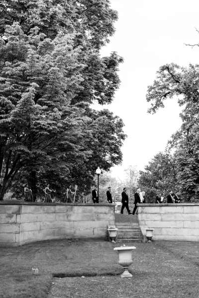 Wedding party walking in a candid moment along a garden walkway, groomsmen in tuxedos, with stone wall, trees, and a lamp post in the background.