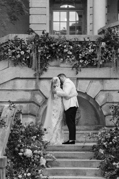 Wedding kiss portrait of a couple kissing on steps, her lace gown trailing as they embrace against a grand stone staircase and wall.