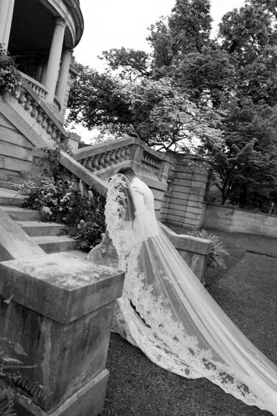 Couple portrait of a bride and groom on stairs, her lace veil trailing on a stone staircase beneath historic columns and garden trees.