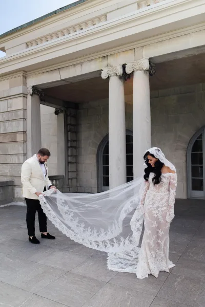 Couple portrait with the groom adjusting the bride's cathedral veil beside classical stone columns and arched windows on a terrace.