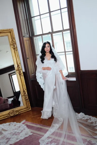 Bridal portrait of a bride in robe with a long lace veil, standing by a tall window with wood paneling and a pink area rug below.