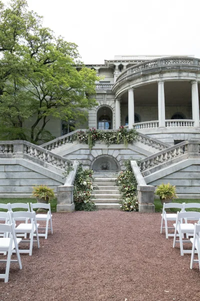 Ceremony setup with outdoor ceremony decor, white folding chairs flanking a gravel aisle before a stone staircase and mansion facade.