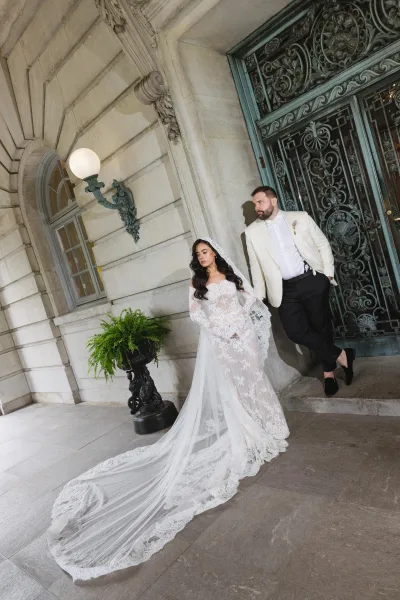 Couple portrait with a bride in lace gown beside a groom, framed by an ornate wrought iron gate and stone architecture