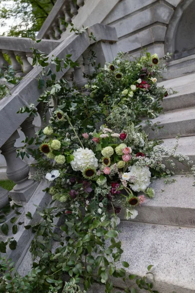 Staircase florals drape as a banister floral garland along a stone balustrade, lush with hydrangeas, framing steps amid garden trees and lawn.