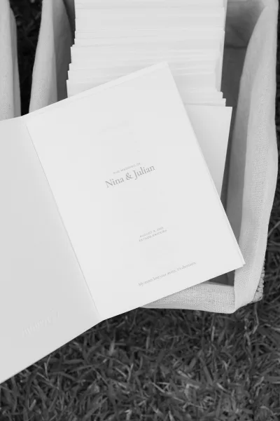 Wedding program with a clean booklet program design, stacked in a linen basket, set on a grass lawn before the ceremony.