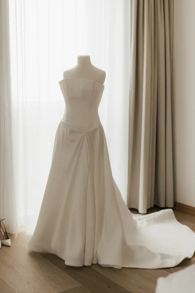 Wedding dress, a strapless wedding gown draped on a dress form, backlit by a window with sheer curtains and beige drapes on a warm wood floor.