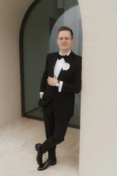 Groom portrait of a black tux groom with a boutonniere, standing by an arched window against a light wall and stone tile floor.