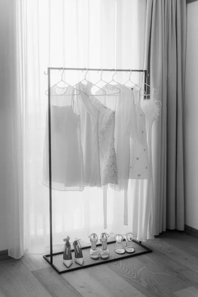 Bridal details featuring a wedding dress hanging on a garment rack, with sheer curtains and a hardwood floor softly lit in the background.
