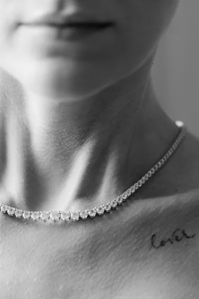 Bridal necklace styled as a classic tennis necklace drapes the collarbone, sparkling beside a delicate love script tattoo.