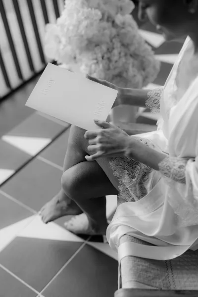 Bride reading a letter in a quiet getting ready moment, seated in a bridal robe near a balcony railing with a simple chair behind.