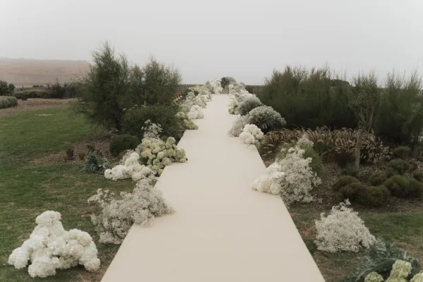 Ceremony aisle design for an outdoor ceremony setup, lined with baby's breath on a grass lawn with distant hills beneath an overcast sky.
