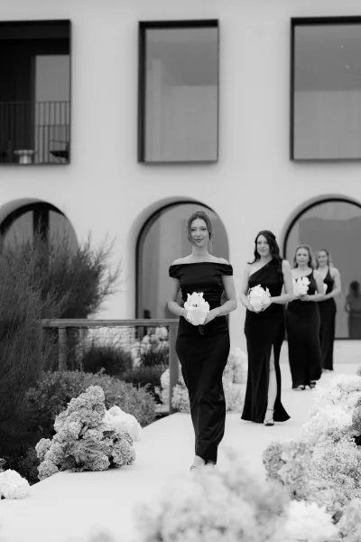 Bridesmaid processional showcasing bridesmaid bouquet ideas as they walk past aisle flowers beside a modern facade with arched windows.