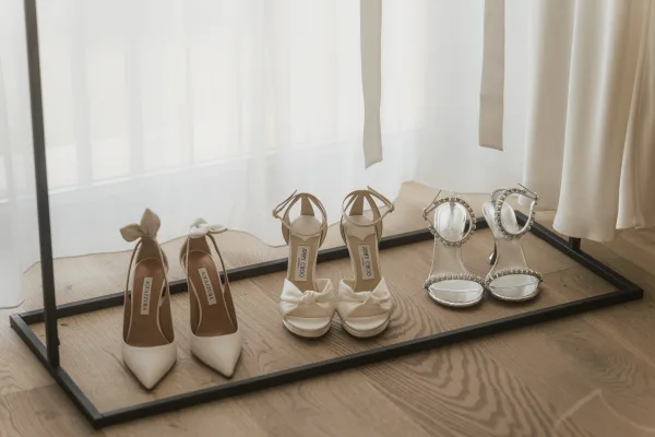 Bridal shoes, from ivory bridal pumps to sleek heels, displayed by a sheer curtain on a wood floor, with subtle crystals catching light