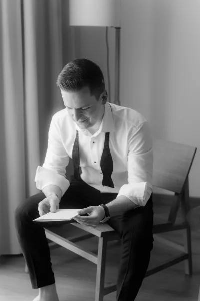 Groom reading letter during a groom getting ready moment, seated indoors with a bow tie at his collar, absorbed in the words.