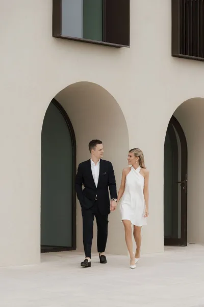 Couple portrait of a bride and groom walking hand in hand beneath arched stucco doorways, her halter-neck dress swaying.
