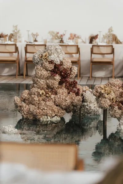 Wedding floral installation forms a floating floral display over pool water beside a long dining table, with hydrangeas softening the scene.