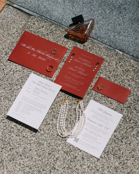 Invitation suite with burgundy wedding invitations arranged in a flat lay with rings on a terrazzo stone surface, plus perfume and pearl jewelry