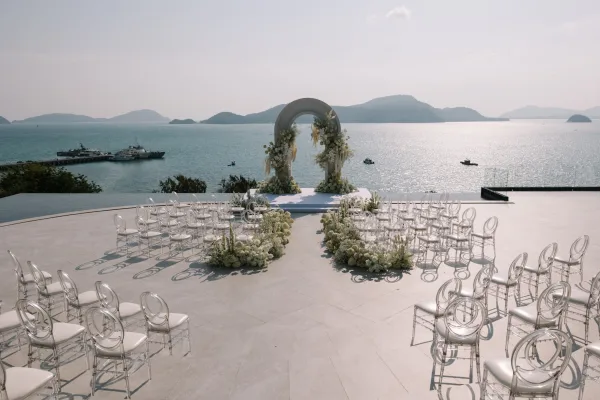Ceremony setup for an outdoor wedding ceremony with a white floral arch and clear chairs on a terrace overlooking the ocean and islands