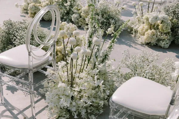 Ceremony aisle decor with ground flower aisle of white hydrangeas and baby’s breath, lining acrylic chairs on a stone patio outdoors