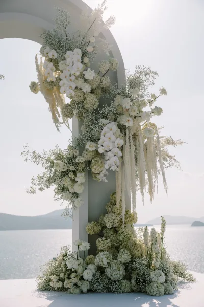 Wedding ceremony arch with white floral arrangement and pampas accents, set oceanfront with distant mountains and sunlit sky behind