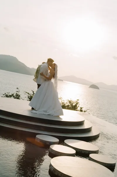 Wedding kiss portrait at sunset with bride and groom kissing, her veil blowing beside an infinity pool terrace with ocean and mountains behind
