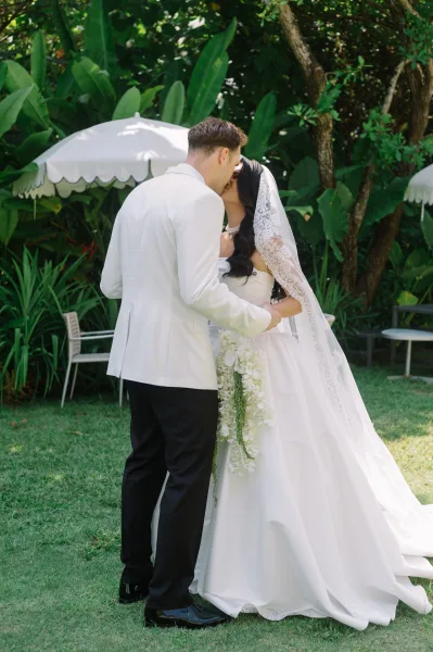 Wedding kiss as bride and groom embrace, her lace veil and cascading white bouquet visible on a tropical lawn with patio umbrellas behind