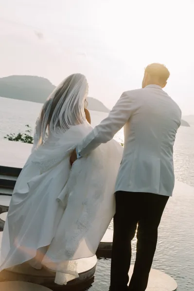 Couple portrait from behind of bride in a strapless gown and veil with lace train beside groom in white jacket at sunset terrace above ocean