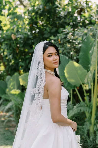 Bridal portrait of a bride looking back in a strapless wedding dress, holding a bouquet with a lace veil against lush tropical greenery backdrop