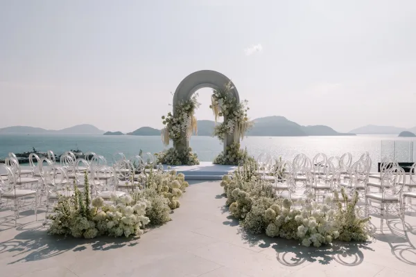 Ceremony setup for an outdoor wedding ceremony with a white floral arch, clear chairs, and aisle florals on a terrace overlooking ocean and mountains