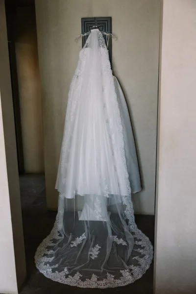 Wedding dress with lace wedding dress details, tulle skirt and long train on a hanger by a doorway against a neutral wall