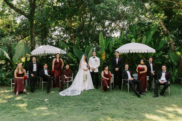 Wedding party portrait with bride and groom, bridesmaids in burgundy dresses and groomsmen in tuxedos, under white umbrellas amid tropical greenery