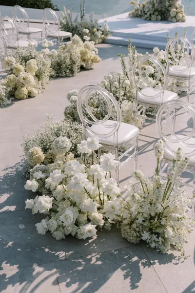 Ceremony aisle decor with wedding aisle flowers lining the stone patio, clear acrylic chairs and a modern stage set against water backdrop