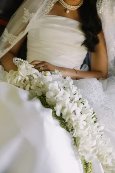 Bridal portrait of a bride in a strapless wedding dress with lace veil, pearl jewelry, and white bouquet, seated indoors on a chair