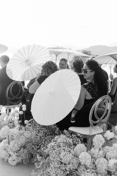 Ceremony guests hold wedding guest parasols and wear sunglasses on clear acrylic chairs beside floral aisle arrangements in mountain air