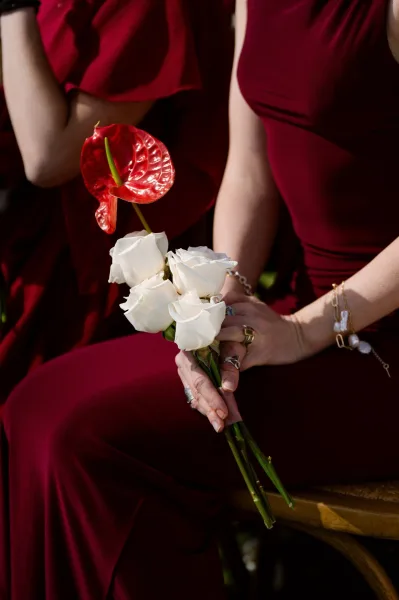 Bouquet close-up of a white rose bouquet with a single red anthurium, set against burgundy fabric draped over a wooden chair.