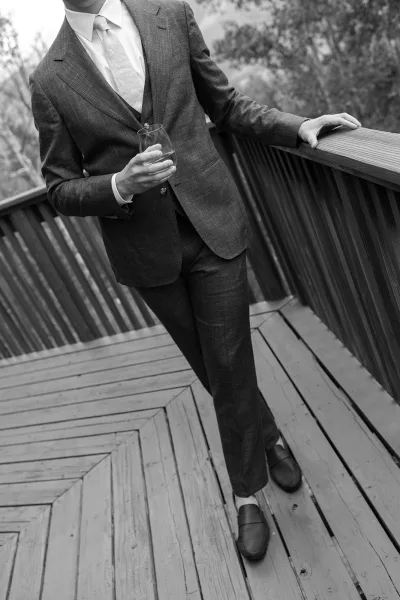 Groom portrait in a three-piece suit, leaning on a deck railing with a whiskey glass, trees behind and loafers visible