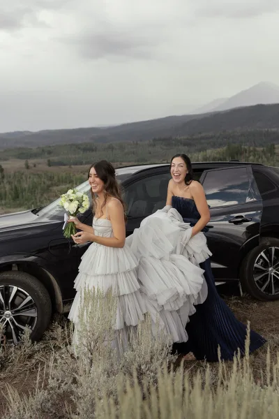 Bride with bridesmaid portrait, bridesmaid holding her ruffle tulle train as the bride holds a white rose bouquet by a car, mountains behind