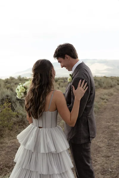 Couple portrait of bride and groom holding a white rose bouquet on a dirt path, mountains behind; her tea-length dress shows a ring