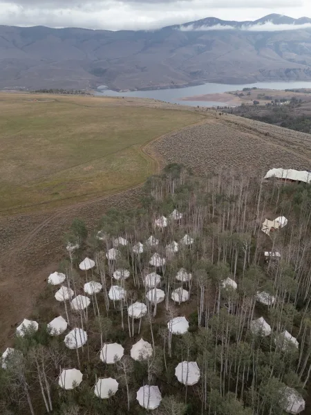 Wedding tented camp with white canvas tents and a large event tent in an aspen meadow, with lake and mountains under cloudy sky