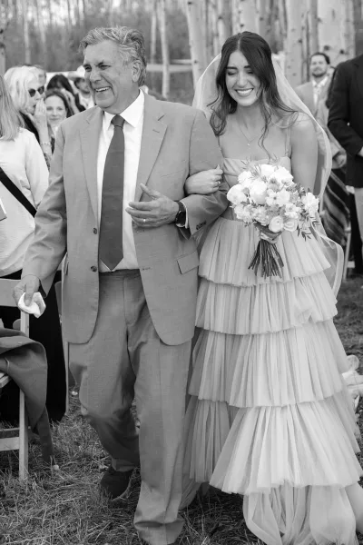 Bridal processional as bride walking down aisle with her father, smiling in tiered tulle gown holding bouquet at outdoor ceremony chairs