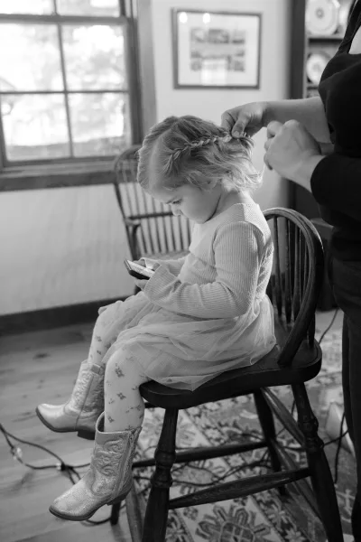 Flower girl getting ready with braided hair, sitting on a wooden chair in a tulle dress and cowboy boots, looking at a smartphone by window light